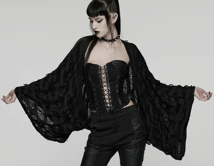 Goth model in black open-knit draped cardigan with wide sleeves over a lace-up corset and black pants.